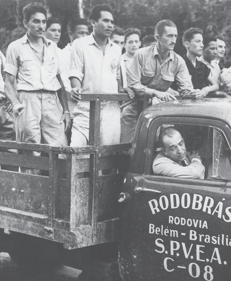 Fotografia em preto e branco. Diversos homens em cima da carroceria de um pequeno caminhão. Na parte da frente há um homem dentro do caminhão com o braço apoiado na janela do passageiro. Os homens que estão na carroceria olham para a frente. Na porta da frente do caminhão, está escrito: Rodobrás. Rodovia Belém Brasília.