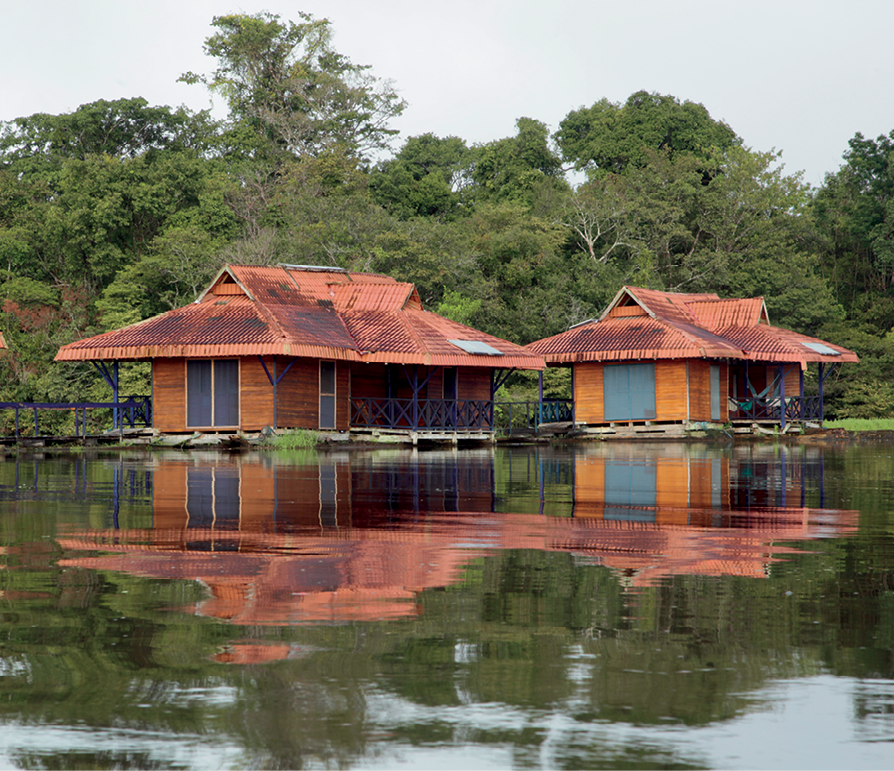 Fotografia. No primeiro plano, vista de um rio com duas edificações flutuantes construídas em madeira, cobertas com telhado vermelho e com varandas ao redor acompanhadas de parapeitos em azul. No segundo plano, na parte de trás das construções, vegetação verde e densa.
