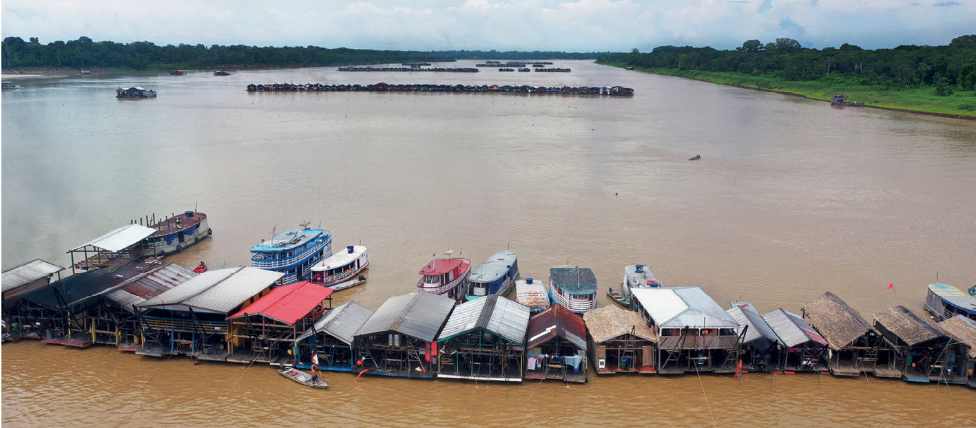 Fotografia. Vista de um extenso rio com diversas construções baixas ao longo de seu curso, dispostas no sentido horizontal. Há alguns barcos parados na frente das construções. Nas margens direita e esquerda do rio, vegetação densa.