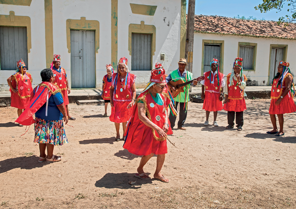 Fotografia. Vista de um grupo de pessoas vestidas com tecidos vermelhos decorados e adereços coloridos na cabeça. Entre elas está um homem segurando um violão. Ao fundo, construções antigas com portas e janelas.