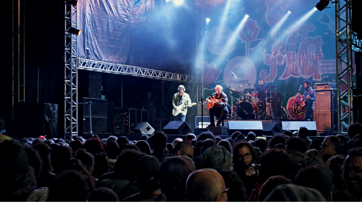 Fotografia. Vista de um palco onde estão quatro pessoas, cada uma tocando um instrumento musical (um violão, uma guitarra, um contrabaixo e uma bateria). Elas estão fazendo uma apresentação para o público que se encontra de frente para o palco, observando os músicos.