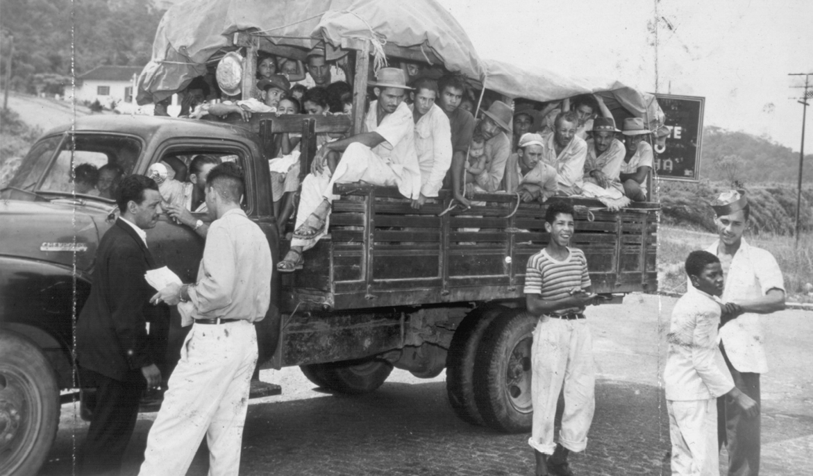 Fotografia em preto e branco.  Um pequeno caminhão com muitas pessoas tentando se acomodar na parte de trás, em cima da carroceria. Ao redor do caminhão estão outras pessoas em pé.