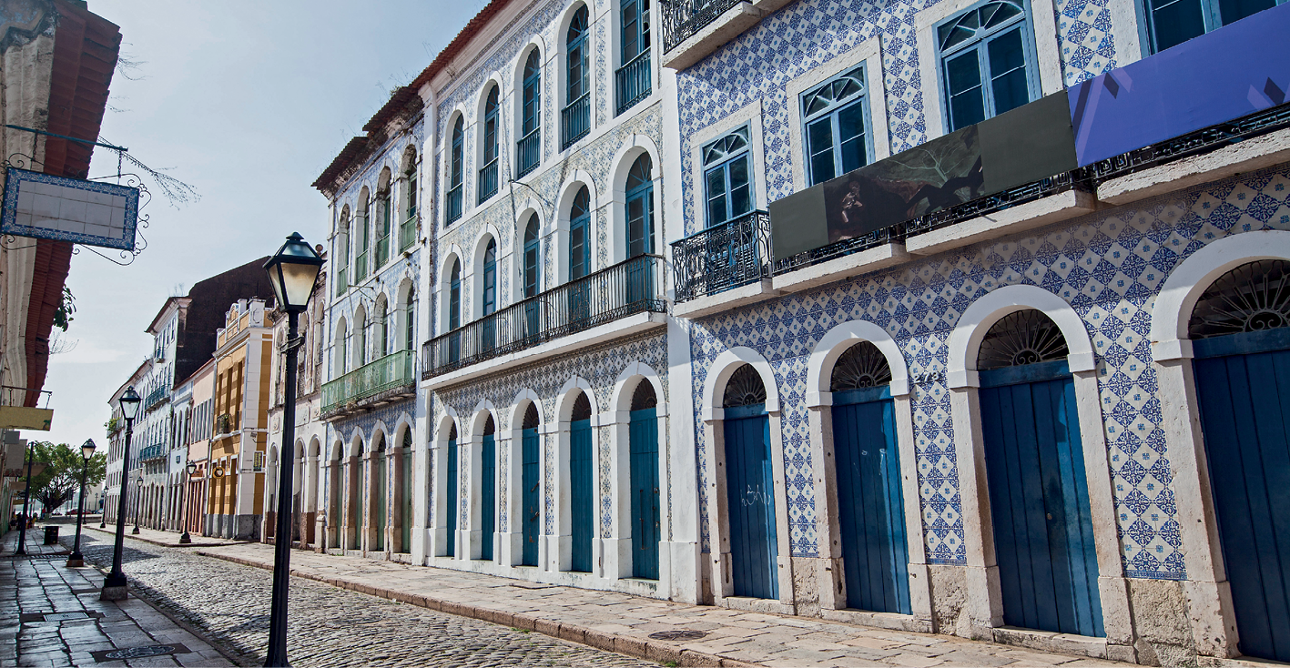 Fotografia. Construções vistas de frente. Elas são altas e possuem muitas janelas e portas em formato de arco. À frente, uma pequena rua de pedras.