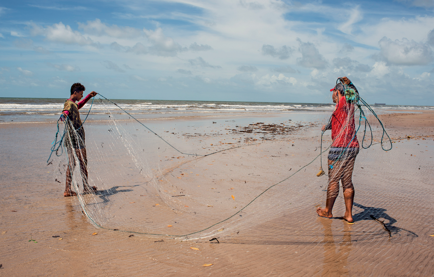Fotografia. Vista de uma praia com uma extensa faixa de areia e a água do mar ao fundo. Em destaque, na areia, dois homens manuseando uma rede de pesca.