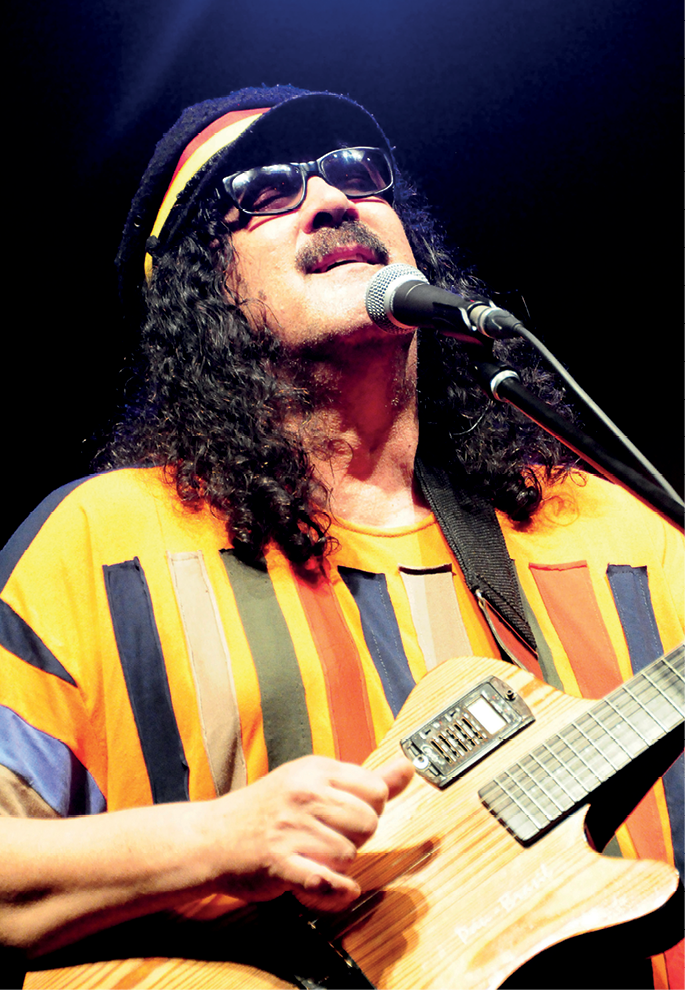 Fotografia. Um homem de bigode, com cabelos pretos e cacheados na altura dos ombros. Ele usa óculos de sol e chapéu e segura uma guitarra e está na frente de um microfone.