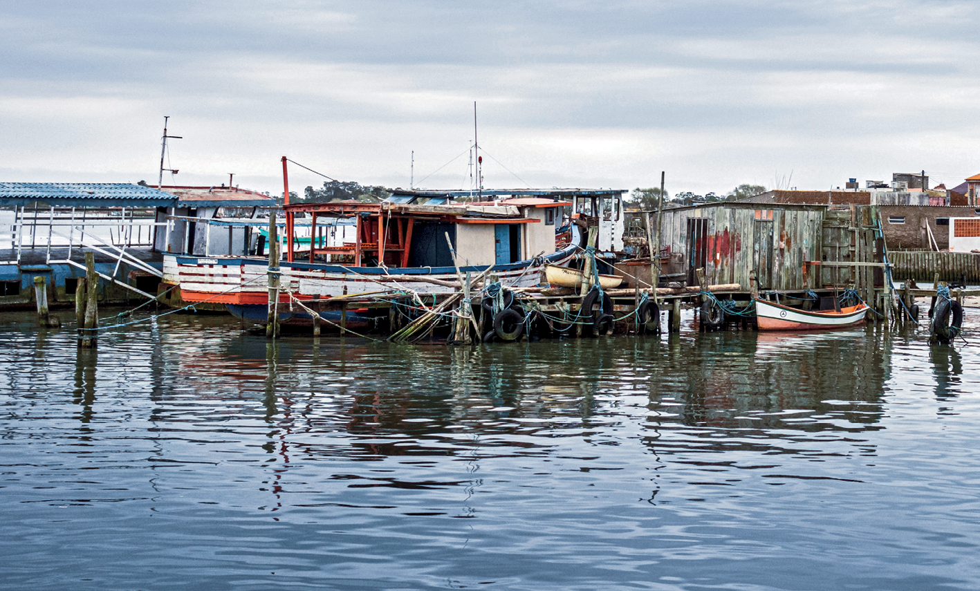 Fotografia. Vista de algumas embarcações de pesca na água e ancoradas próximas a um pier. Ao fundo, algumas construções feitas de madeira.