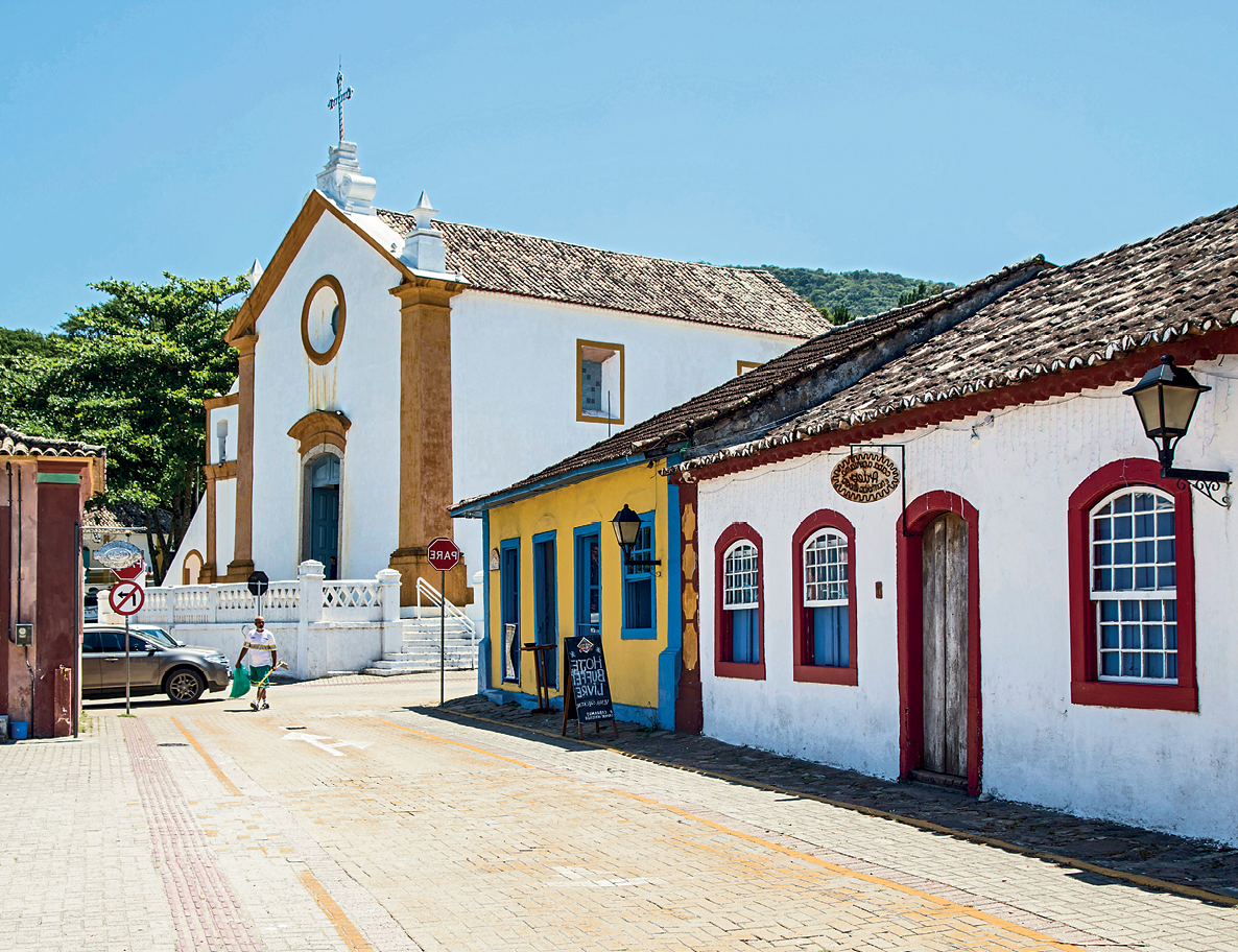 Fotografia A. Vista de uma rua estreita e dois casarios antigos à direita, com janelas amplas, portas junto a rua e telhados inclinados para frente. Ao fundo, no final da rua, uma igreja com uma cruz no topo do telhado.