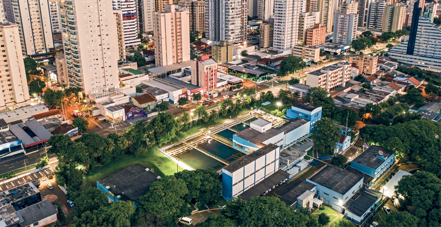 Fotografia. Vista do alto de algumas quadras de uma cidade, com ruas e avenidas se cruzando e uma concentração de edifícios altos, de vários andares, na parte de cima da imagem. Na parte de baixo, em destaque, uma estação de tratamento de água, com alguns prédios e tanques usados para o tratamento da água. Muitas árvores no estorno da estação de tratamento de água.