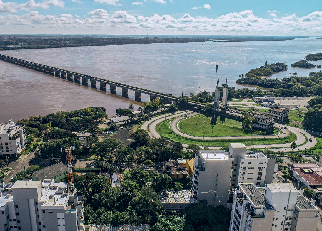 Fotografia. No primeiro plano, vista do alto de parte de uma cidade com prédios, árvores e uma rotatória conectada a uma ponte que atravessa um rio caudaloso. No segundo plano, vista da outra margem do rio, a partir de onde se estende uma vasta área de relevo aplainado.