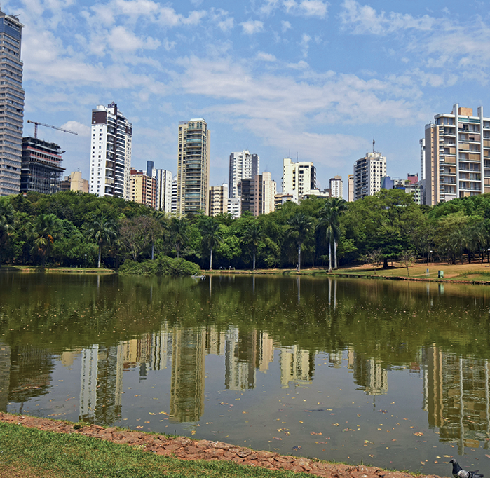 Fotografia. Em primeiro plano, vista de um parque com um lago cercado de árvores. Em segundo plano, diversos prédios.