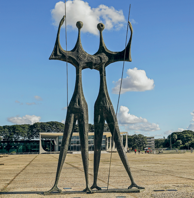 Fotografia. Vista de uma grande escultura de duas pessoas com corpos finos e cabeças pequenas, em cor cinza. Elas seguram uma vara fina e longa, que está posicionada ao lado do corpo. Ao fundo, um prédio de vidro e pilares brancos, além de árvores.