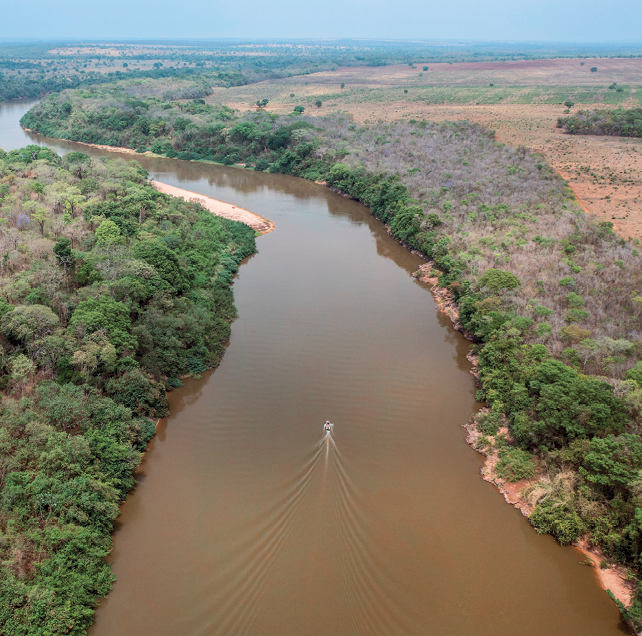 Fotografia. Vista de um extenso rio, ao centro, com vegetação densa formada por árvores em suas margens. Na parte direita da imagem, ao fundo e mais distante da margem, a vegetação é rasteira, há árvores dispersas e uma parte do solo está exposta. Uma pequena embarcação passa pelo rio.