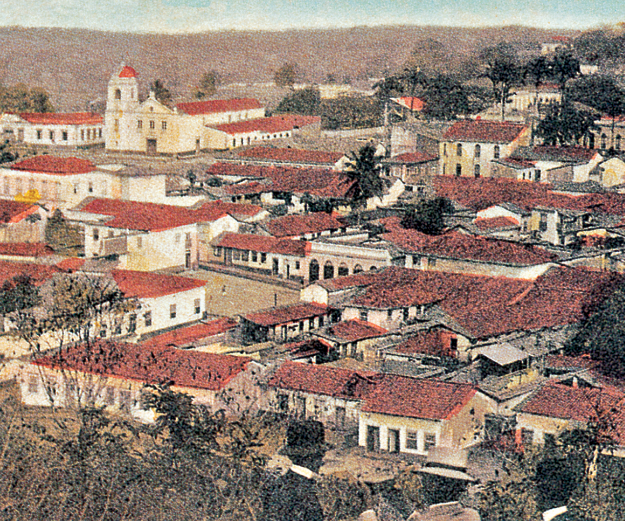 Fotografia. Vista de casas térreas com paredes brancas e cobertas com telhas vermelhas. Ao fundo, há uma igreja. No horizonte, morro. Há algumas árvores nas ruas. No primeiro plano, dois homens e uma mulher de costas e com chapéus.