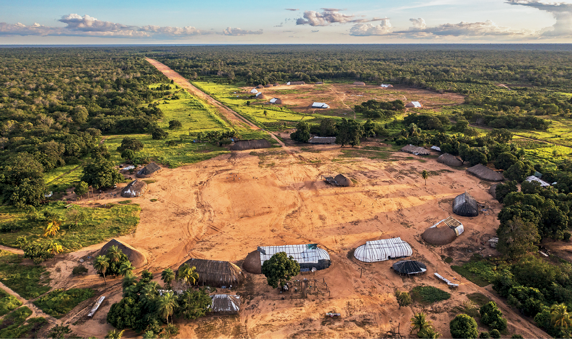 Fotografia. Vista do alto de uma vasta área de relevo aplainado. No primeiro plano, em espaço em formato de círculo com chão de terra batida; na parte de cima, outro espaço em formato circular e com as mesmas características. No terceiro plano, vista de uma extensa área de vegetação densa. Uma longa estrada de terra corta a floresta, ligando-se a esses espaços com construções.