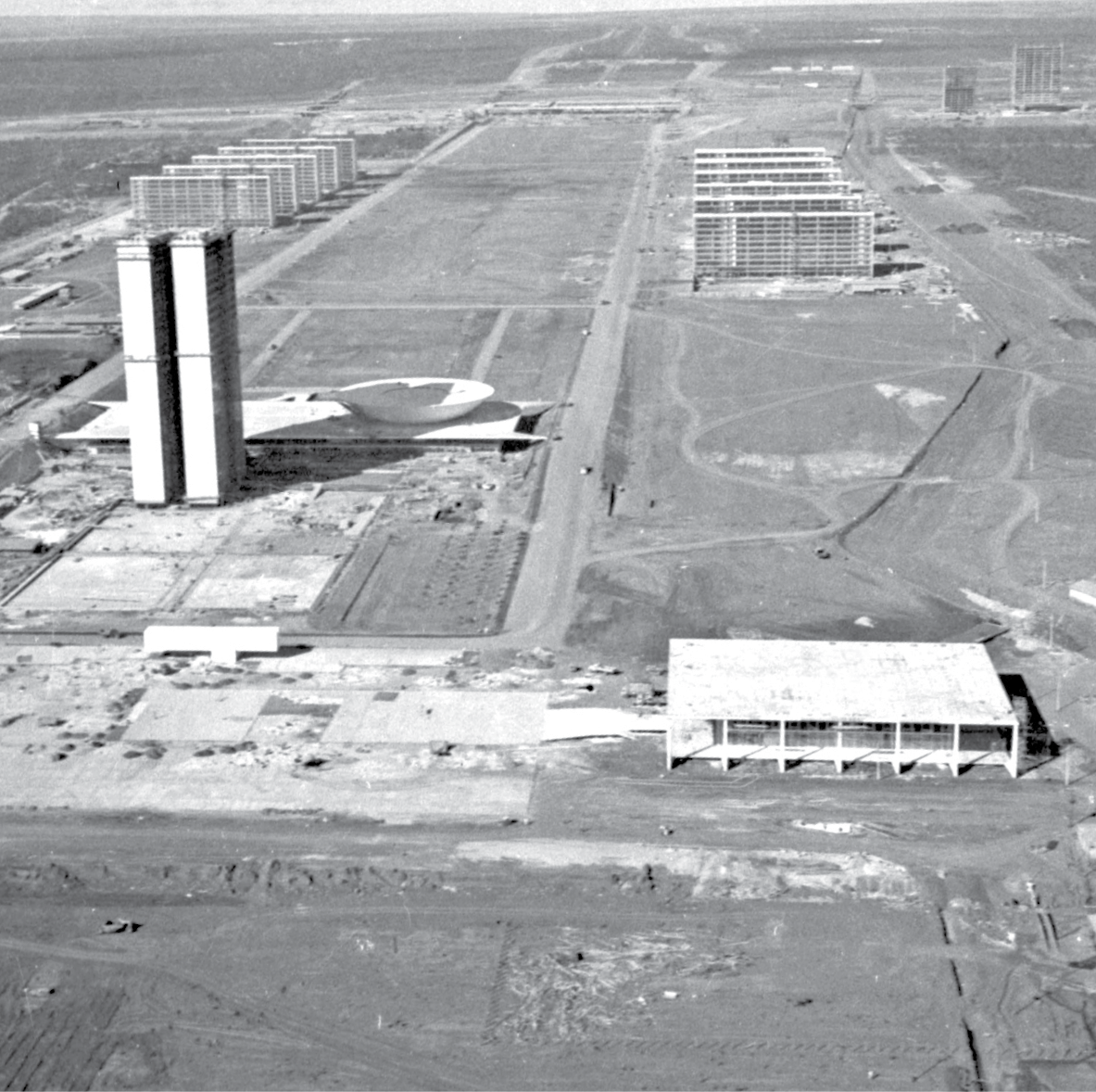 Fotografia em preto e branco. No primeiro plano, à direita, vista de um prédio em formato retangular, com diversas pilastras no entorno. À esquerda, dois edifícios altos, de vários andares, idênticos, alinhados lado a lado. Atrás deles, uma construção em formato de cúpula côncava. No segundo plano, um largo canteiro com duas vias sendo construídas paralelamente, à esquerda e à direita. Ao lado de cada via, diversos prédios enfileirados. Os prédios são retangulares e com vários andares construídos.