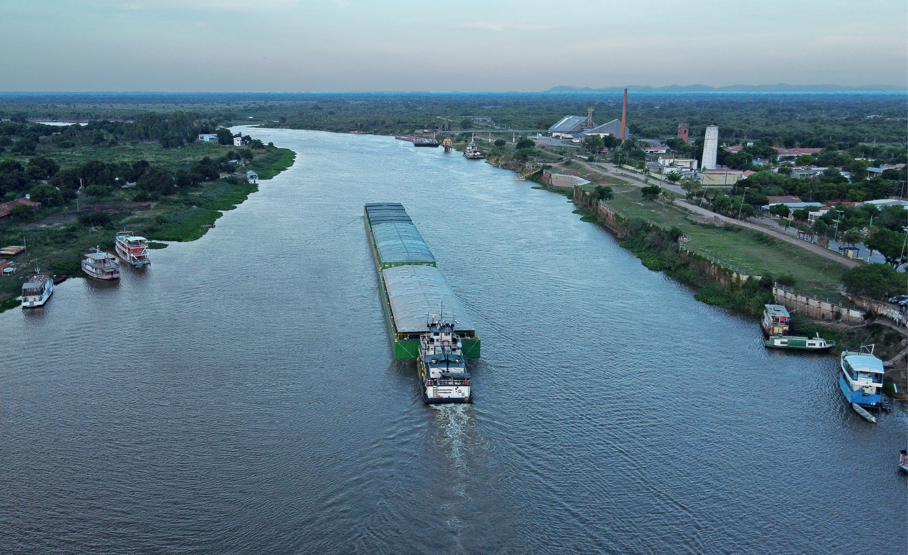 Fotografia. No primeiro plano, vista de um rio largo com uma grande embarcação navegando no meio do canal. Nas margens, barcos ancorados e algumas construções. No segundo plano, vista de uma extensa área de vegetação.