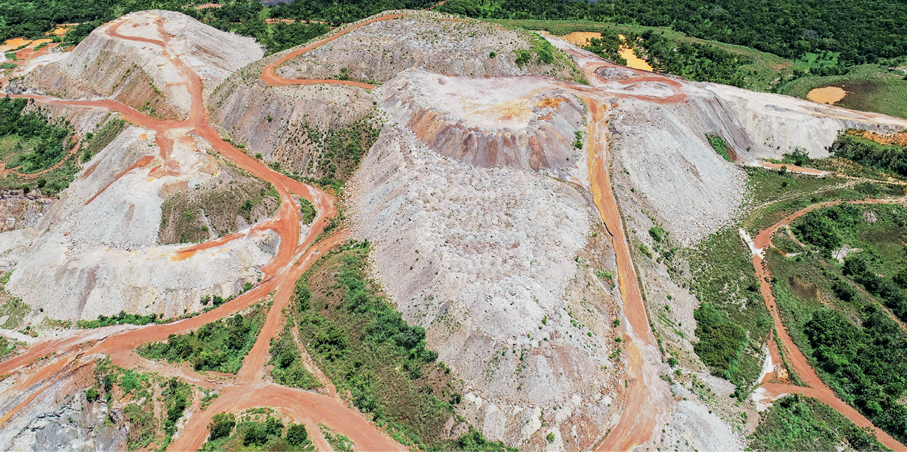 Fotografia. Vista do alto mostrando uma área com grandes montes de material particulado, como areia e cascalho, gerado pela atividade de mineração. Entre os montes e ao redor deles, há estradas de terra e pequenas lagoas na parte de cima.