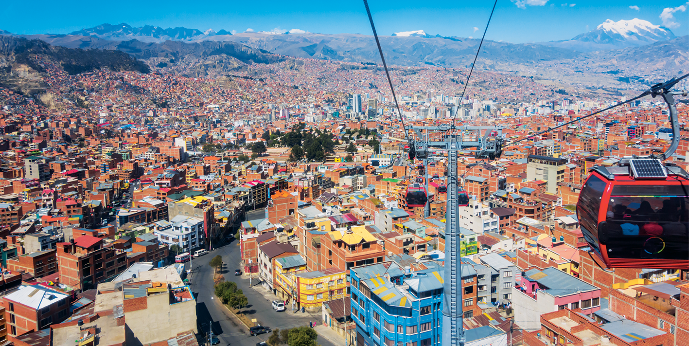 Fotografia. Vista de uma grande cidade com muitas construções em tonalidade marrom. Ao redor da área urbana há montanhas. As construções são próximas umas das outras. Há ruas asfaltadas e poucas árvores. Ao fundo, uma das montanhas está com neve no topo.