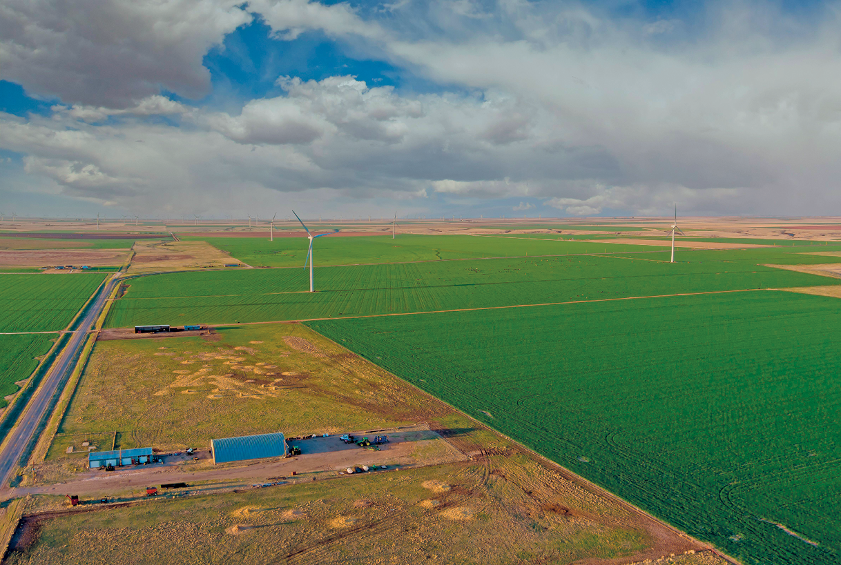 Fotografia. Vista de uma extensa área com plantação à direita e ao fundo. Há duas hélices de geração de energia eólica na plantação. Na parte esquerda da foto, há uma rodovia pavimentada, galpões e área sem vegetação.