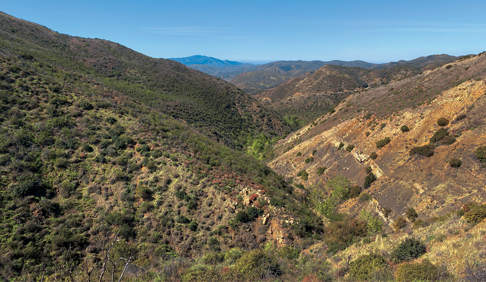 Fotografia. Vista de um vale ao centro da fotografia e montanhas com vegetação baixa e arbustiva. Há pontos sem vegetação, com solo exposto. Céu azul.