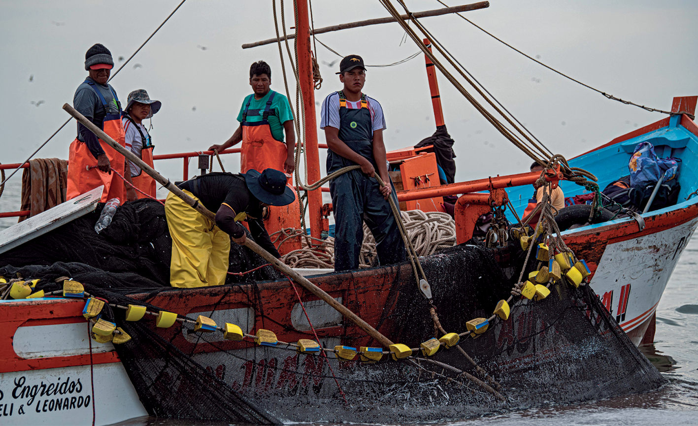 Fotografia. Fotografia de uma pequena embarcação de pesca com cinco tripulantes usando macacões azul, amarelo e laranja. Com o uso de uma haste grande, um tripulante manipula a rede na lateral do barco enquanto outro a segura com uma corda. Ao fundo, os outros três observam.