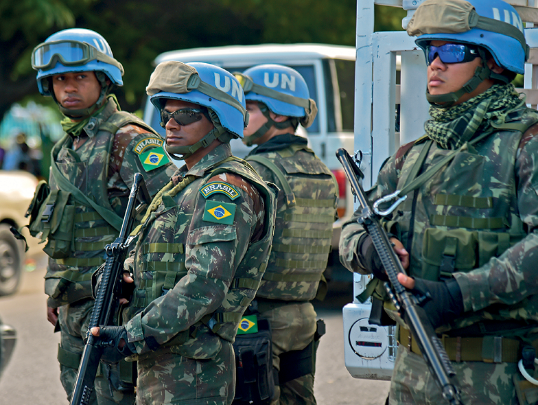 Fotografia.  Um grupo de quatro soldados em pé, segurando fuzis, usando roupas militares com a bandeira do Brasil estampada na parte superior da manga do casaco, capacetes azuis com a sigla "UN", óculos escuros.