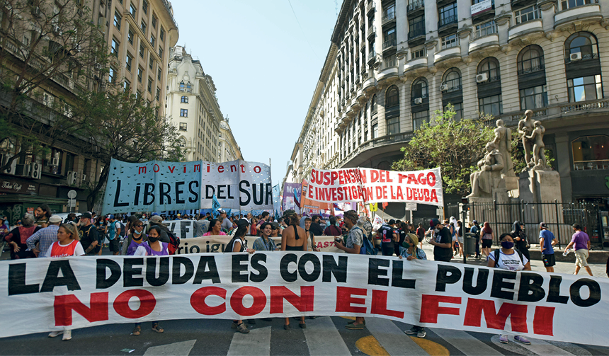 Fotografia. Em uma rua, atrás da faixa de pedestres, pessoas alinhadas horizontalmente seguram uma faixa com os dizeres: "La Deuda es con el pueblo. No con el FMI". Atrás dessas pessoas, uma multidão segue com outros cartazes e faixas, ocupando o asfalto. Nas laterais, prédios altos de arquitetura neoclássica.