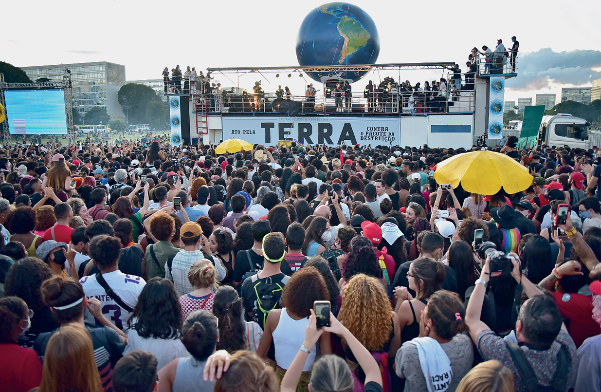 Fotografia. Multidão aglomerada de frente para um trio elétrico ao fundo, com diversas pessoas no palco, onde há uma faixa com a palavra "TERRA". Atrás do trio, no alto, um grande balão redondo  com o desenho dos continentes, representando o globo terrestre. Ao fundo, céu entre nuvens e topos de prédios mais altos.