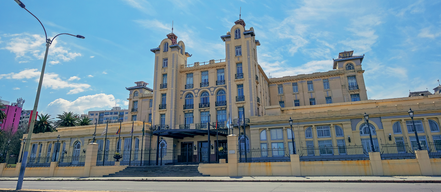 Fotografia. Vista da fachada de uma construção antiga com muitas janelas e portas grandes. A parte da frente da construção é térrea e a parte de trás tem quatro pavimentos. No centro da construção, duas torres paralelas separadas por área de quatro pavimentos. À esquerda, ao fundo, topo de outros  edifícios mais modernos.