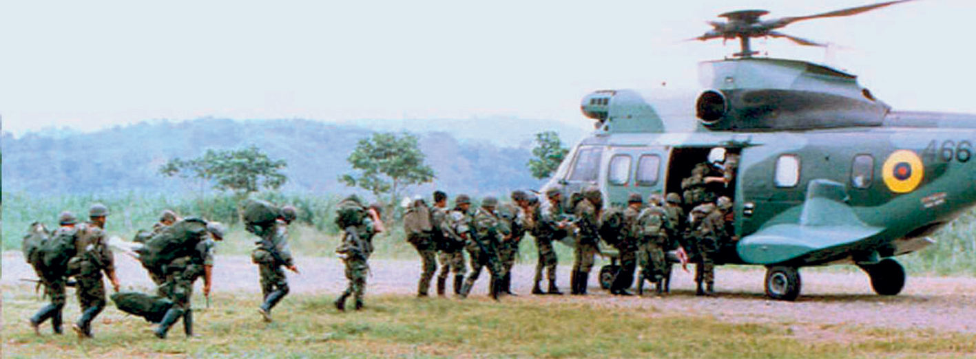 Fotografia.  Em uma área plana, aberta, soldados enfileirados da esquerda para a direita, em direção a um helicóptero militar, que está com a porta lateral aberta para a entrada dos soldados, que carregam mochilas e sacolas militares. Ao redor, vegetação.