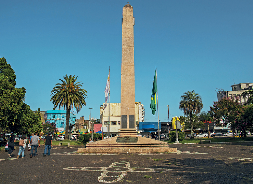 Fotografia. Vista de uma praça de asfalto com destaque para um obelisco de concreto alto e retangular. Ao seu redor, dois mastros com bandeiras. À esquerda, pessoas caminhando. Ao fundo, área urbanizada e árvore isoladas.