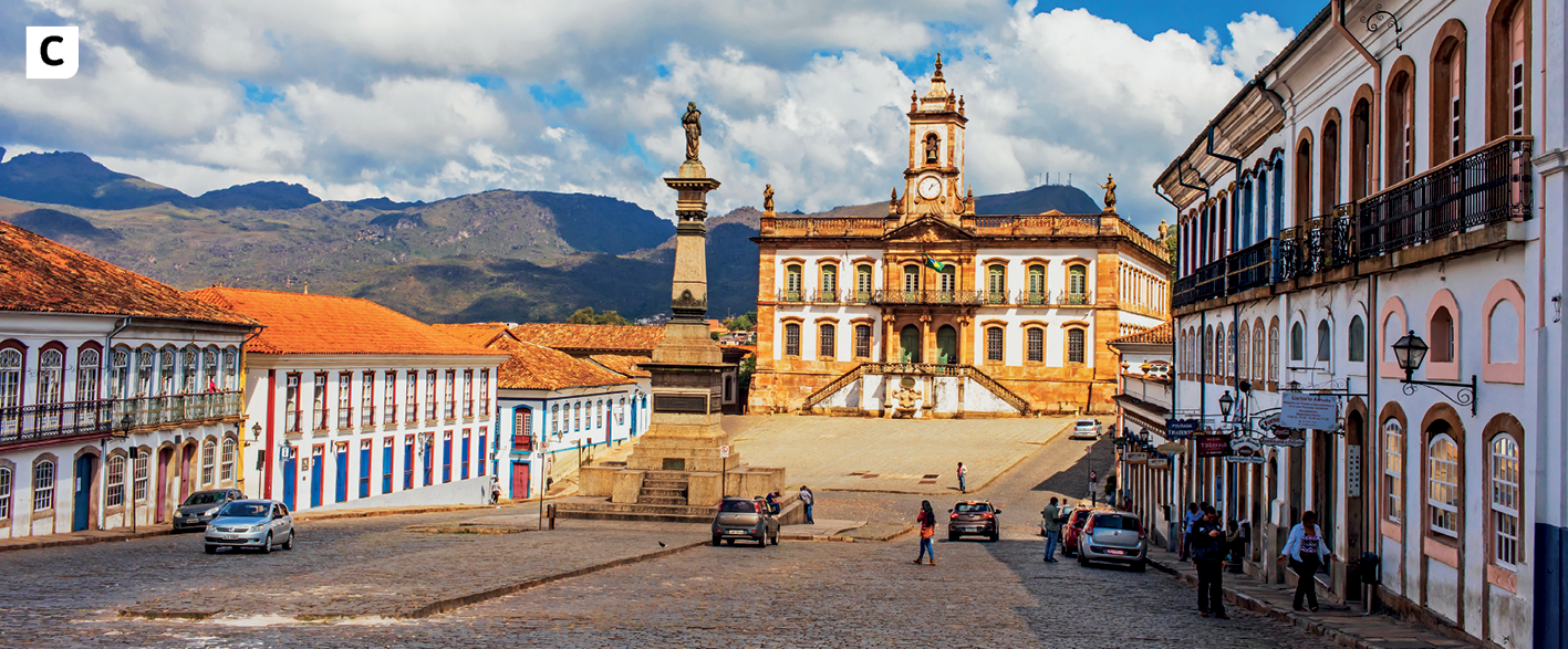 Fotografia. Vista para rua  larga, de paralelepípedos, com poucos carros e algumas pessoas caminhando nas calçadas. Nas laterais da rua,  casarões de arquitetura barroca, um ao lado do outro, com muitas janelas e portas grandes. Depois dos casarões, uma praça com um monumento alto e uma estátua no topo e uma igreja com uma torre no meio, diversas portas e janelas nos dois pavimentos. Ao fundo, montanhas e o céu azul com muitas nuvens brancas.