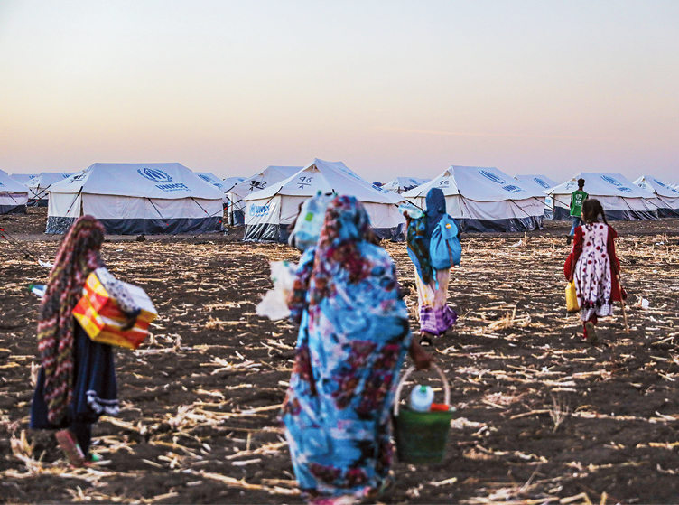 Fotografia. No primeiro plano, grupo de mulheres de costas em pé sobre um campo com solo escuro, sem vegetação e em parte coberto por resíduos no chão que lembram palha. No segundo plano, várias tendas brancas usadas para abrigar pessoas refugiadas.
