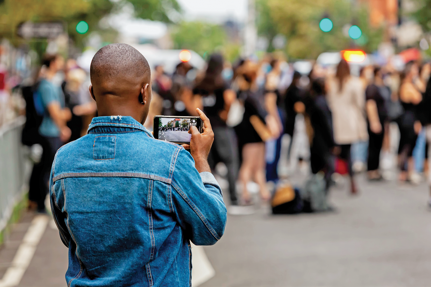 Fotografia. No primeiro plano, uma pessoa vista de costas da cintura para cima. Ela segura um celular na horizontal com a câmera aberta. À frente dela, diversas pessoas com a imagem desfocada na rua.
