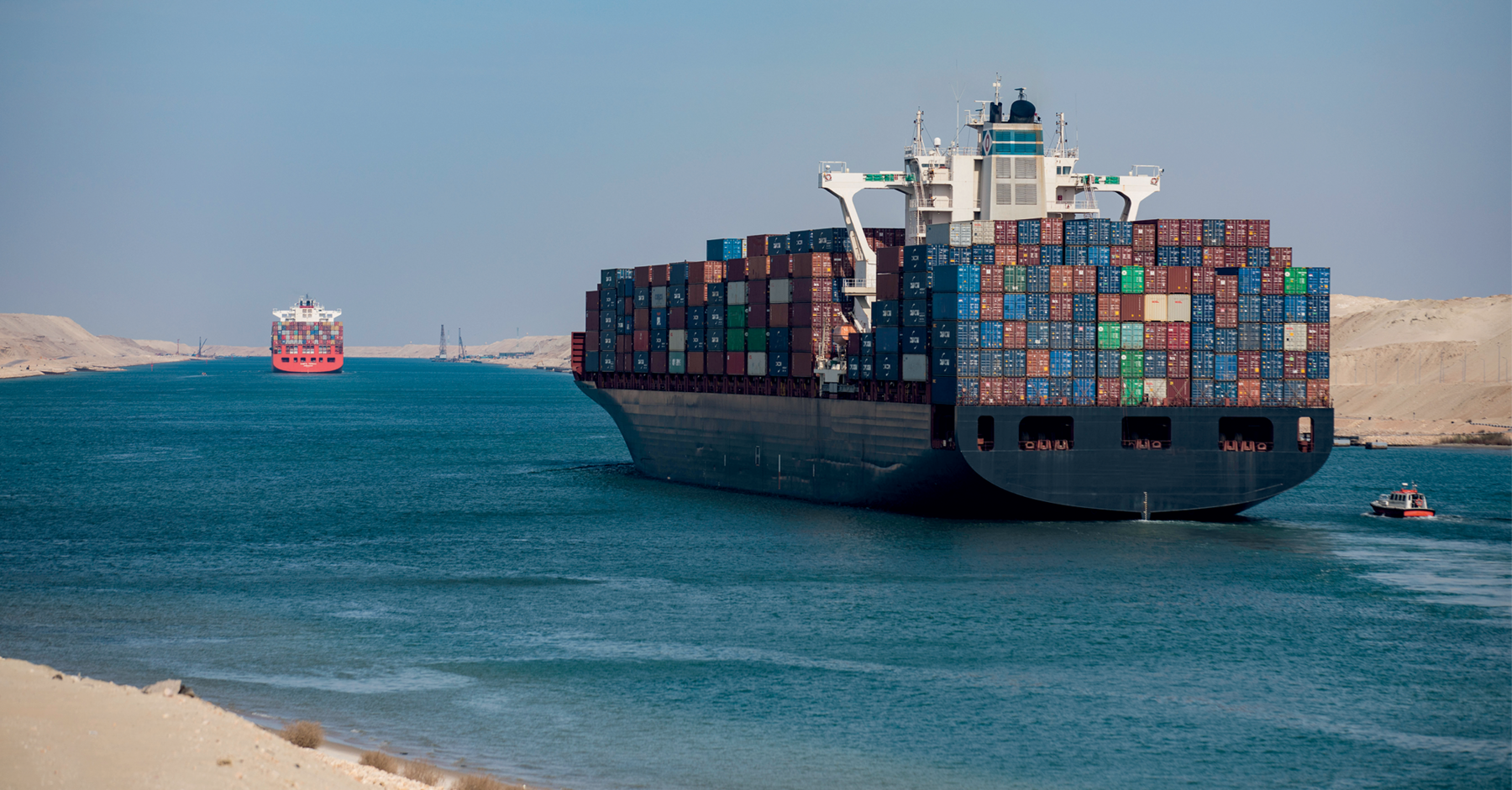 Fotografia de um navio sobre as águas de um grande canal. Há centenas de containers no navio. Ao fundo. um segundo navio também grande. Ao redor do canal por onde passam os navios estão pequenas montanhas de areia. Ao lado do primeiro navio, um pequeneo barco.