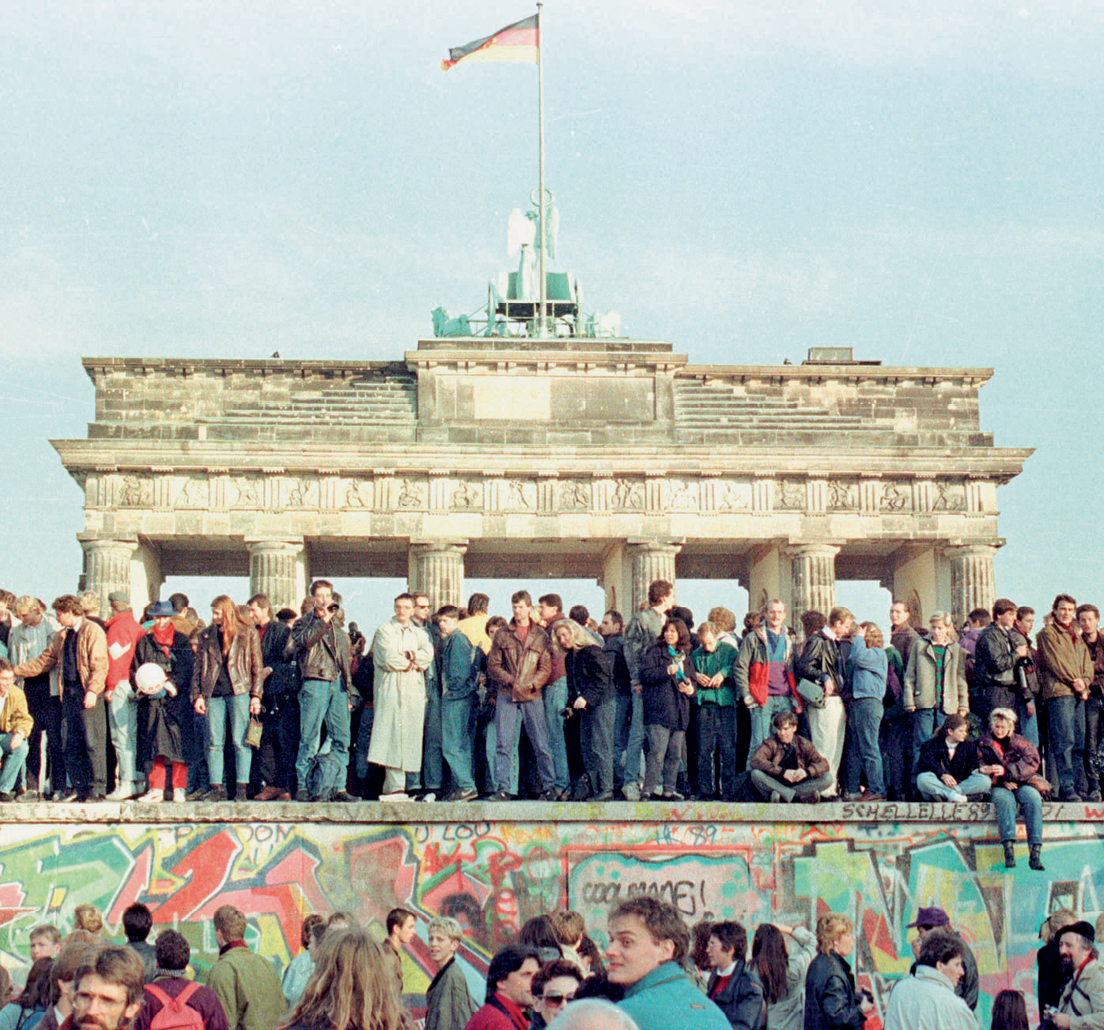 Fotografia. Vista de um grupo grande pessoas em pé em cima de um muro e, atrás deles, um portal feito com pilares e uma torre com a bandeira da Alemanha. No muro há escritos coloridos. Na frente do muro, no primeiro plano da foto, também há pessoas em pé.