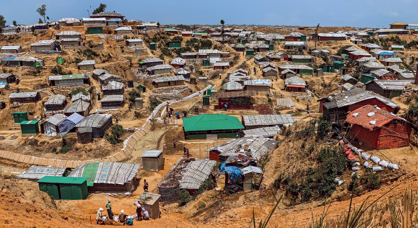 Fotografia. Vista de uma área de superfície irregular, com o solo exposto e muitos abrigos feitos de bambus, lonas e outros materiais, dispostos de forma irregular no terreno. Entre os abrigos, nas ruelas que se formaram, algumas pessoas reunidas em grupos e outras circulando. 