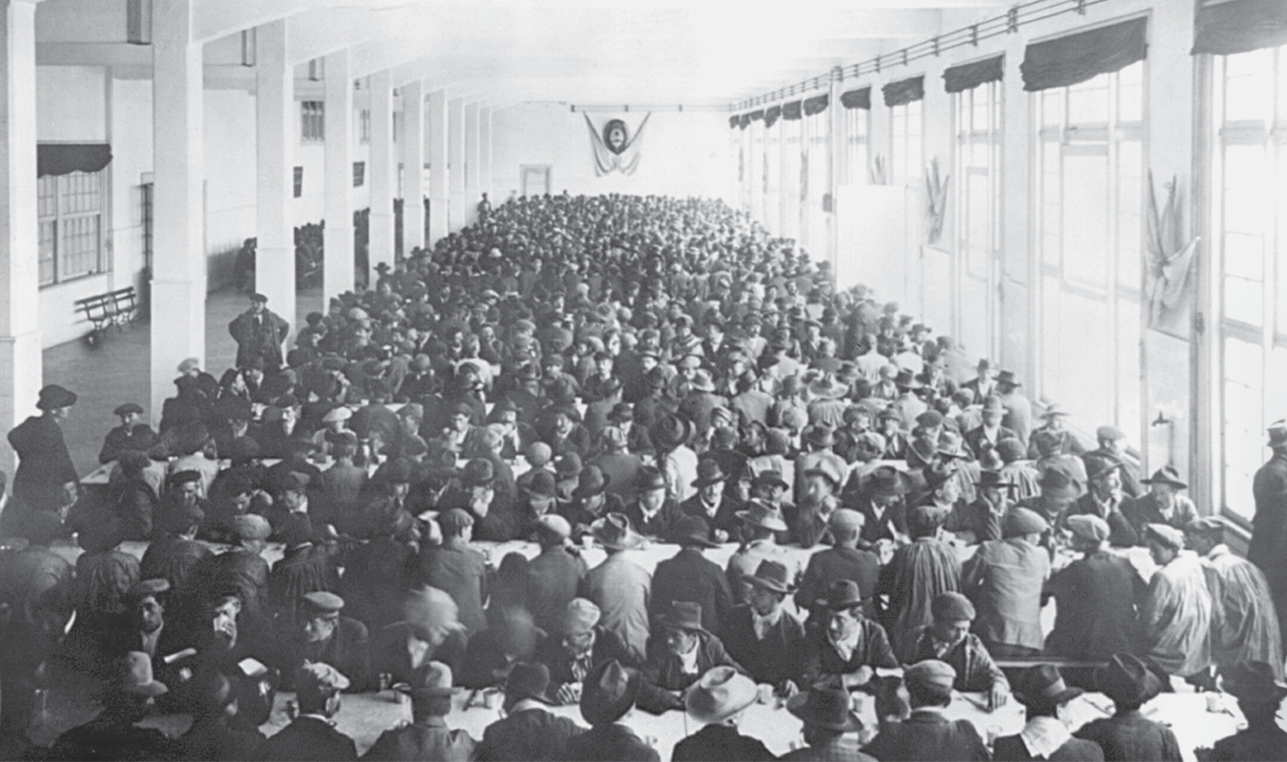 Fotografia em preto e branco. Vista de um salão amplo, com diversas janelas à direita e uma fileira de colunas à esquerda. Nesse espaço, um grande número de pessoas reunidas. Elas estão sentadas em bancos junto a mesas compridas e dispostas paralelamente, ocupando todo o salão.