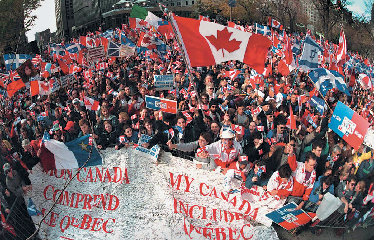 Fotografia. 
Vista de um espaço aberto com uma aglomeração de pessoas segurando bandeiras do Canadá, entre outras, cartazes e uma grande faixa branca com escritos em vermelho no primeiro plano.