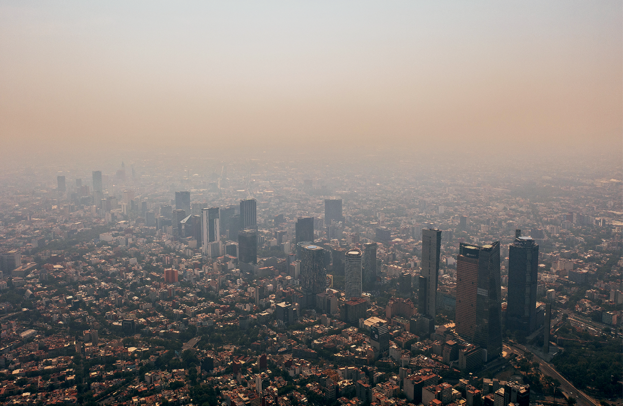 Fotografia. Vista de uma grande cidade. Na parte central, diversos edifícios altos se destacam na paisagem, com residências e prédios menores no entorno. No céu, é perceptível uma camada densa de gases e poluentes em suspensão de cor alaranjada.