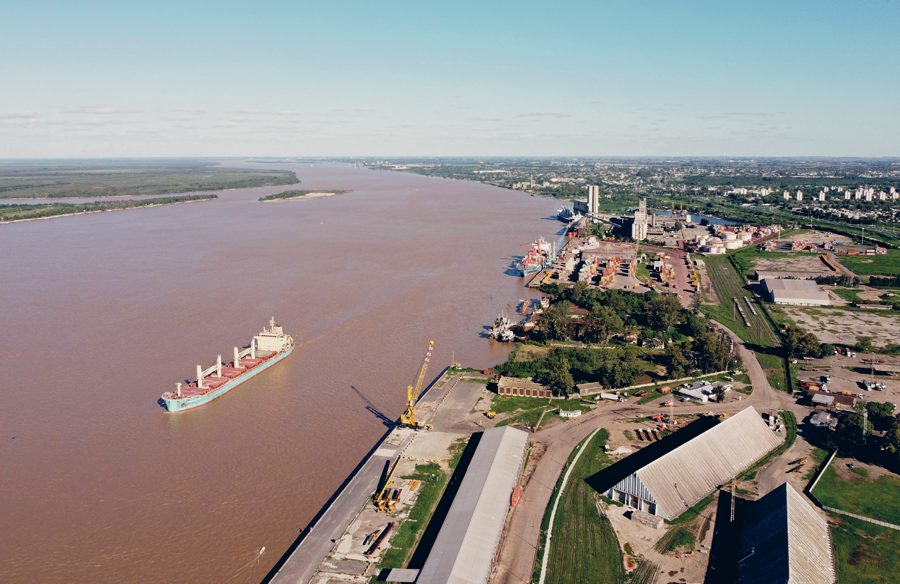 Fotografia. Vista de uma cidade às margens de um rio. À esquerda da foto, há o rio com grande largura entre as margens e água em tom marrom, com um navio cargueiro. À direita, na margem, há uma plataforma cimentada e uma área mais ao fundo cimentada e com vários contêineres. Um navio cargueiro está atracado nessa margem. Mais à direita, galpões, algumas áreas com solo exposto e pequenas construções. Ao fundo, área urbanizada com a presença de muitas áreas verdes. Ao fundo, o horizonte com o rio.