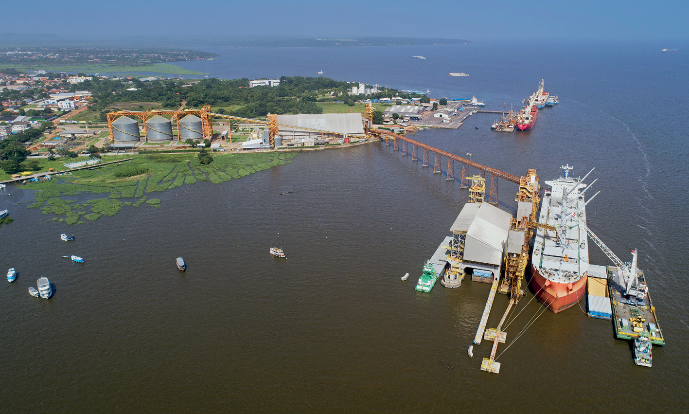 Fotografia.  Vista de uma extensão de água, muito larga e de tonalidade escura, à direita e no primeiro plano da foto. No segundo plano, área de terra emersa com três grandes silos interligados por uma estrutura laranja que avança pela água e chega a um navio atracado com uma plataforma ao lado e alguns barcos. Em terra firme, também há vias de acesso, construções e uma área com vegetação densa. No rio há outros barcos navegando e outro navio atracado mais ao fundo. Ao fundo, o horizonte do rio e o céu nublado.