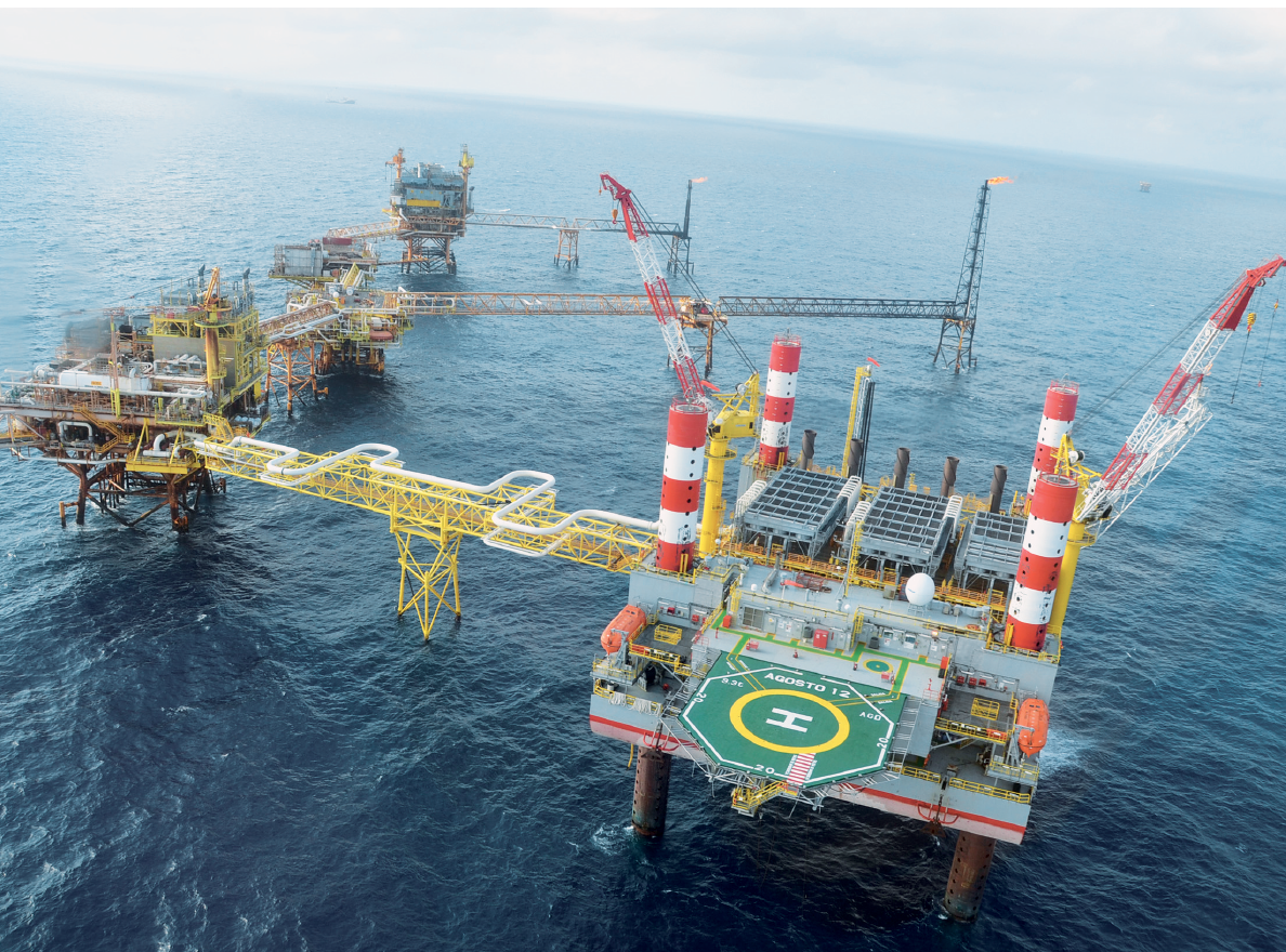 Fotografia. Vista de uma grande plataforma de exploração de petróleo sobre o mar. A estrutura está suspensa por pilares de concreto. Há tubos na vertical, uma pista de pouso de helicóptero e três estruturas construídas sobre a plataforma. Uma outra estrutura de metal, que lembra uma ponte, liga a plataforma a uma segunda estrutura de metal suspensa. Ao fundo, dois tipos de estrutura vertical de metal no mar, ligados às estruturas suspensas. No topo das torres há chamas de fogo.