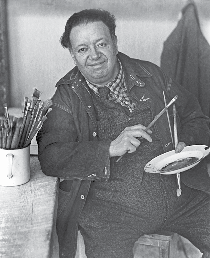 Fotografia em preto e branco. Foto do pintor Diego Rivera. Um homem está sentado em um banco e aparece do joelho para cima. Ele segura um pincel na mão direita e um prato branco com tinta escura na mão esquerda. Ele usa calça, blusa, camisa xadrez, gravata e casaco escuros. Tem cabelos curtos e pretos e está sorrindo. Ao seu lado, uma mesa de madeira, onde ele apoia o cotovelo. Na mesa há uma xícara com vários pincéis. Ao fundo, uma parede com uma sobretudo pendurado.