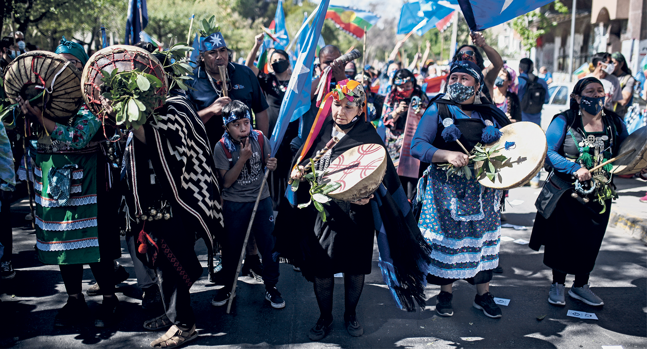 Fotografia.  Em uma área aberta, um grupo de pessoas reunidas performa uma atividade tradicional indígena Mapuche. No primeiro plano, pessoas carregando instrumentos redondos com roupas típicas e bandeiras azuis. Atrás, mais pessoas acompanham as pessoas do primeiro plano.