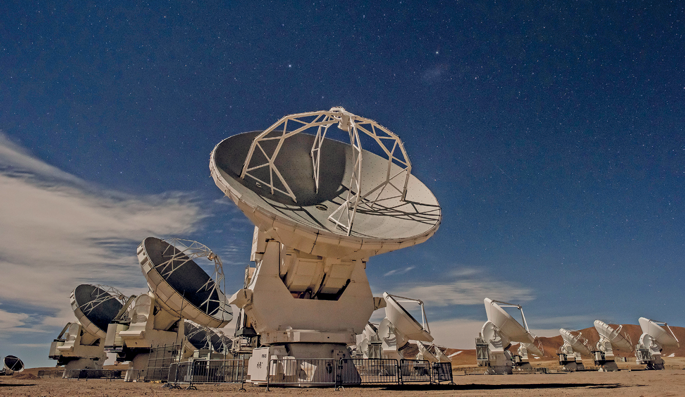 Fotografia. Em uma área aberta, diversos telescópios dispostos em fileiras. No centro da foto, um telescópio e sua base com uma cerca de proteção no solo e a grande estrutura redonda posicionada em direção ao céu.