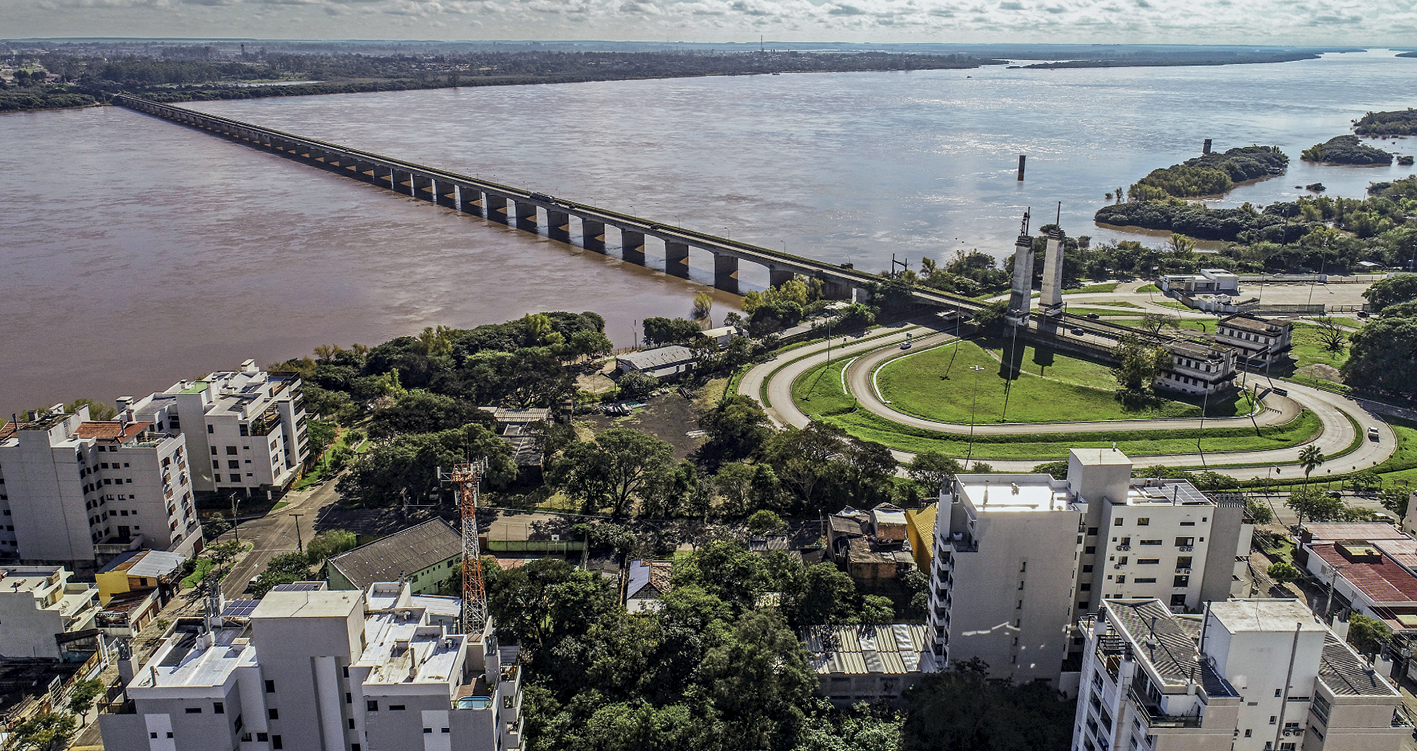 Fotografia. Vista aérea de trecho de rio em tons e marrom em toda a sua extensão, com uma ponte ligando as duas  margens. Na parte inferior da foto, em uma das margens do rio, área urbanizada com prédios, ruas e vias de acesso a ponte, além de árvores e gramados.