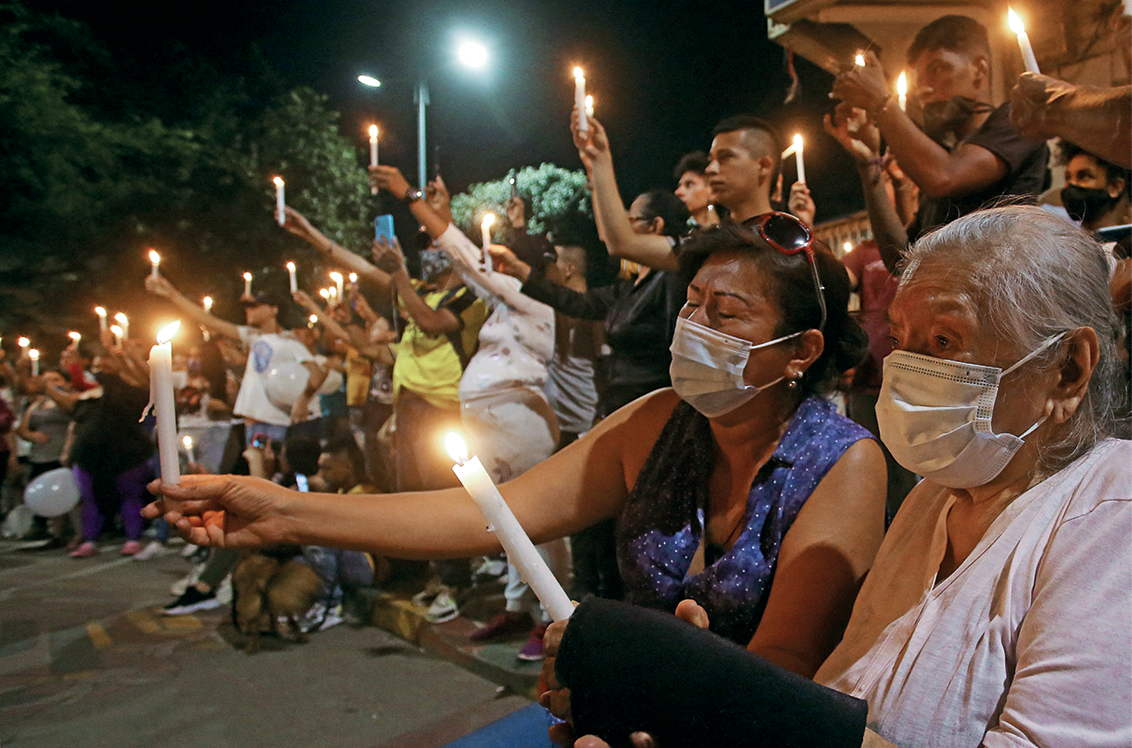 Fotografia. Em uma área aberta e de noite, diversas pessoas reunidas seguram velas acesas. No primeiro plano, duas mulheres idosas e de máscaras de proteção facial seguram uma vela acesa cada. Atrás, jovens em pé e pessoas sentadas também seguram velas com os braços esticados para frente.