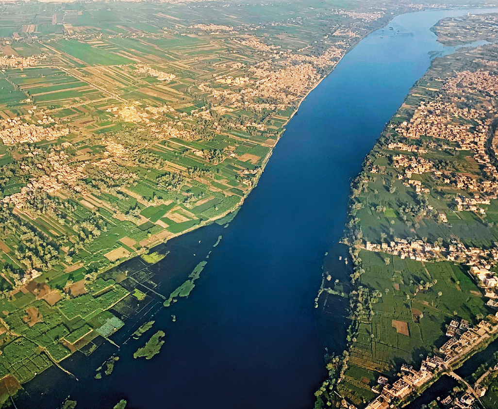 Fotografia. Vista aérea do vale do Rio Nilo. No centro da foto, o leito do rio entre as margens verdes de áreas agrícolas e construções diversas no entorno.