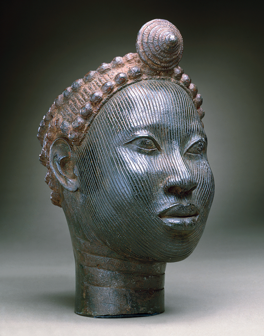 Escultura. Retrato de uma escultura de bronze representando o busto de uma mulher com um adereço no topo da cabeça. A escultura tem tons predominantemente acinzentados.